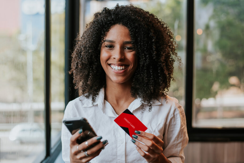 Consumidora segura smartphone e cartão de crédito enquanto sorri para a câmera.