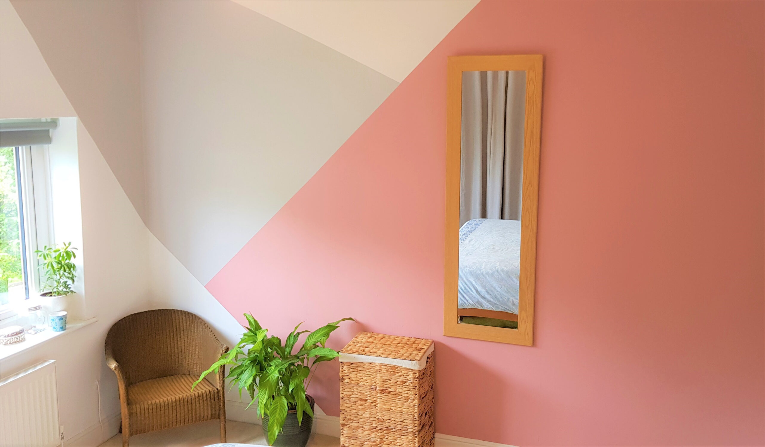 Quarto com paredes pintadas em padrão geométrico, no ambiente vemos uma poltrona, um cesto, um espelho e uma planta. As cores da parede variam entre creme, cinza claro e rosa.