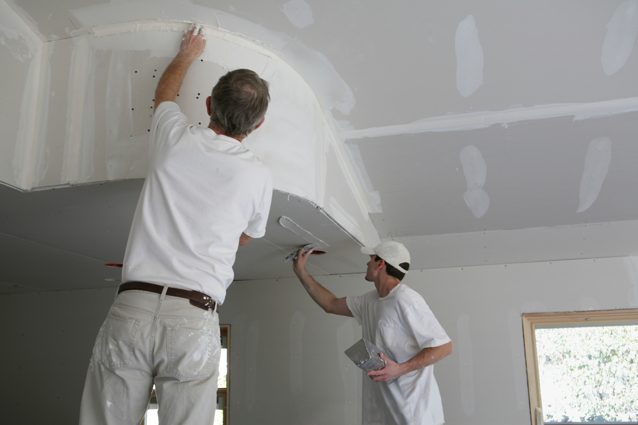 Imagem que demonstra o processo de instalação do drywall, nela há dois profissionais de obra homens, eles são brancos e estão de costas em cima de escadas.     