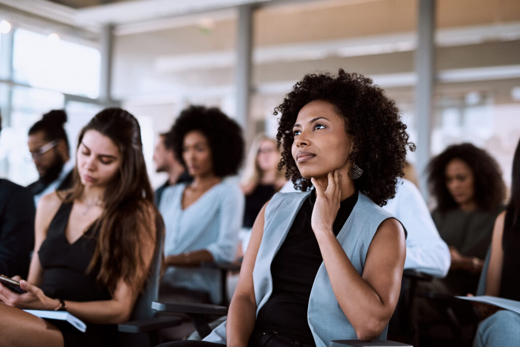 Uma empresária está sentada em uma cadeira durante uma convenção, ela é uma mulher negra por volta dos 40 anos, tem olhar concentrado e a mão apoia o queixo.